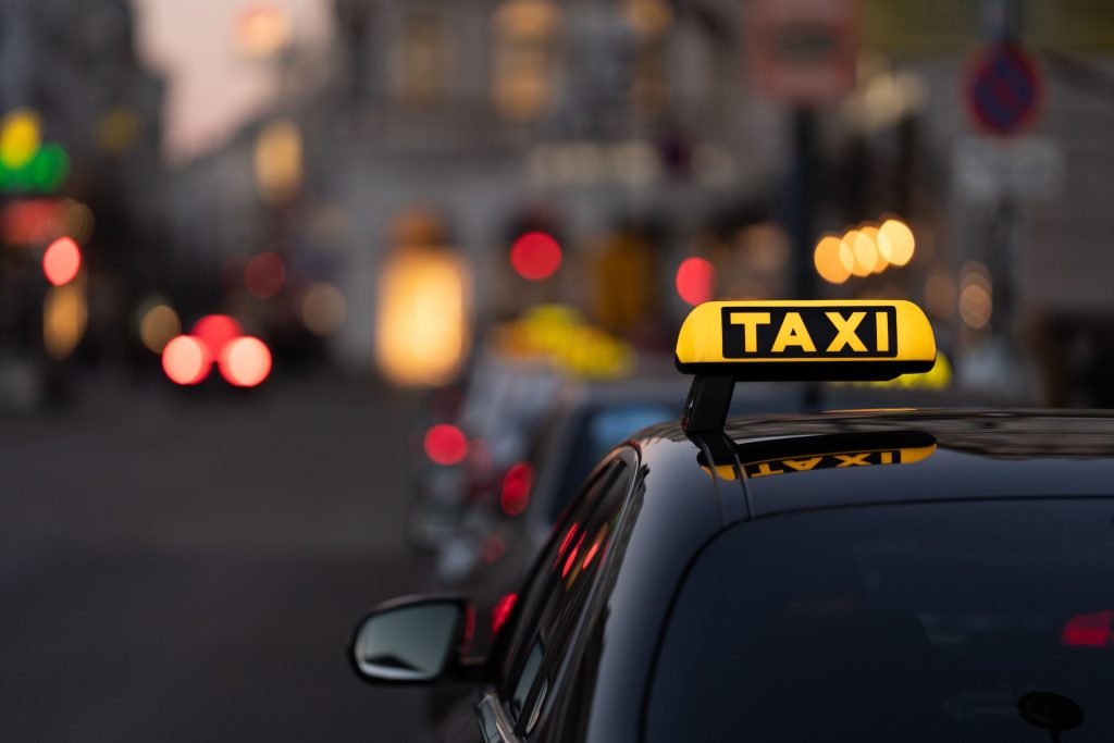 A yellow taxi sign is visible atop the dark, glossy roof of a car traveling on a city street at dusk. The background is blurred, with lights visible from buildings and other vehicles. The overall mood is calm, with an urban setting. This photo could be used for projects about transportation, city life, or travel. The photo could also represent getting around a busy area, or a peaceful night in the city.
