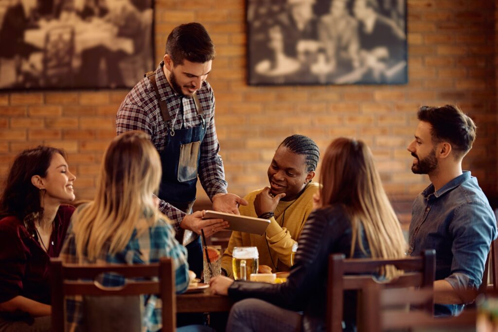 A group of friends are sitting at a wooden table in a restaurant. They are talking to a server who is holding a tablet, presumably showing them the menu.
